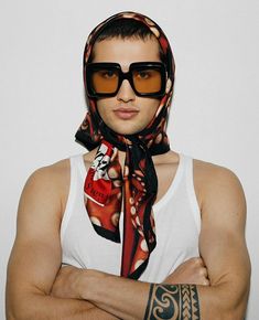 a man with his arms crossed wearing sunglasses and a scarf around his neck, standing in front of a white wall