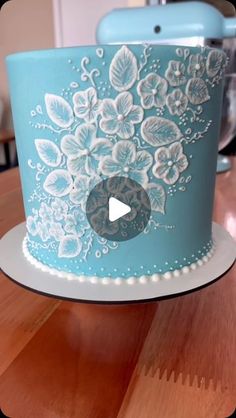 a blue cake with white flowers on it sitting on top of a wooden table next to a blender