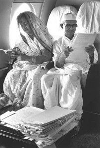 a man and woman sitting on an airplane seat reading papers while looking at the camera