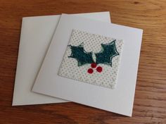 two cards with holly decorations on them sitting on a wooden table next to each other