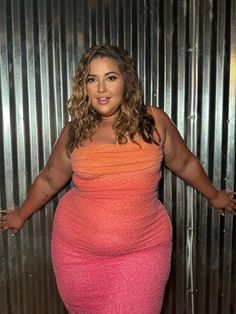 A woman with curly, light brown hair poses confidently in a shimmering ombre dress that transitions from orange to pink, standing in front of a corrugated metal wall.