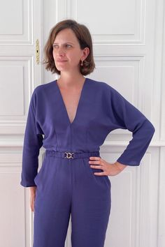 a woman standing in front of a white door wearing a purple jumpsuit with her hands on her hips