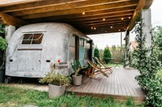 an old camper sits under a covered porch with chairs and potted plants on the deck