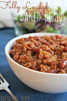 a white bowl filled with baked beans next to a fork and salad on a blue cloth