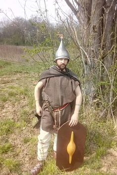 a man dressed in medieval clothing standing next to a tree and holding a plaque with a yellow arrow on it