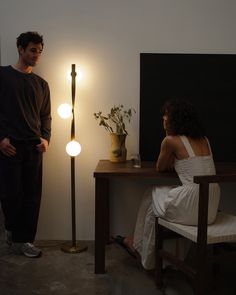 a man standing next to a woman sitting in front of a table with a lamp on it