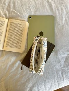 an open book sitting on top of a bed next to an apple laptop and purse