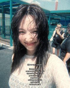 a woman with long black hair standing in front of a fence and people behind her