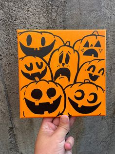 a hand holding up an orange square with black faces on it and pumpkins painted on the sides