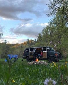 a van parked in the middle of a field with trees and flowers around it on a cloudy day