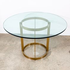 a round glass table with gold metal frame around the base on concrete flooring in front of a white wall