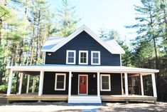 a black house with white trim and red door in the middle of pine trees on a sunny day