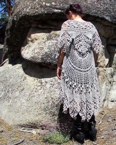 a woman in a crocheted dress standing next to large rocks with grass on the ground