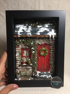 a hand holding up a framed photo of a brick building with red doors and wreaths