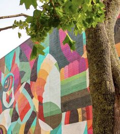 a large colorful mural on the side of a building next to a tree with green leaves