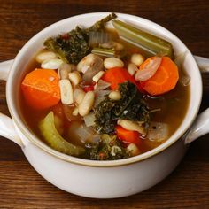 a white bowl filled with soup and vegetables on top of a wooden table next to a spoon