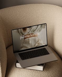 an open laptop computer sitting on top of a book next to a beige chair in a room