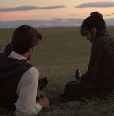 two people sitting on the ground in a field at sunset, one is holding a cell phone