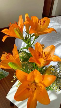 an arrangement of orange lilies in a white vase on a table with greenery