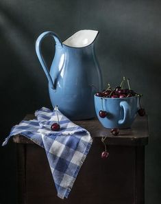 a blue pitcher sitting on top of a table next to a bowl filled with cherries