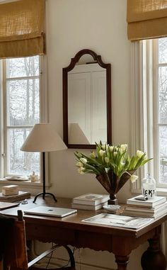 a desk with a mirror, lamp and flowers on it in front of two windows