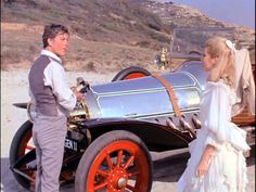 a man standing next to a woman in front of an old fashioned car on the beach