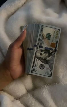 someone is holding cash in their hand on top of a white fluffy blanket with one hundred dollar bill sticking out of it