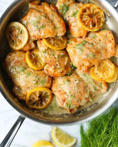 chicken with lemons and herbs in a skillet on a marble countertop next to sliced lemons