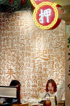 a woman sitting at a desk in front of a wall with chinese writing on it