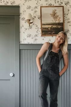 a woman leaning against a wall with her hands on her hips, wearing overalls