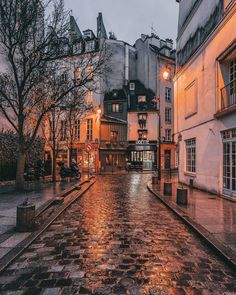 an empty cobblestone street at dusk with buildings in the background and lights on