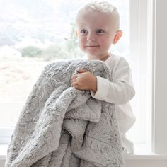 a little boy that is holding a blanket in front of a window with a smile on his face