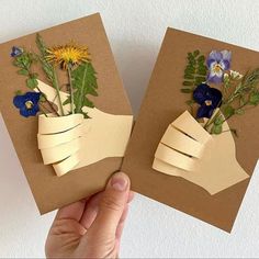 two handmade greeting cards with flowers in vases on brown paper, being held by a person's hand