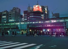 many people are crossing the street in front of some tall buildings at night with bright lights