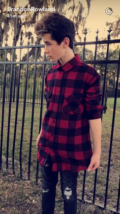 a young man standing in front of a fence wearing black ripped jeans and a red flannel shirt