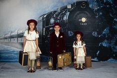 three children are standing in front of a wall with a train on it and suitcases