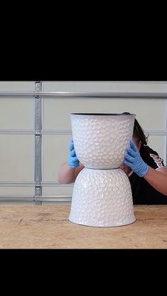 a person with gloves on their hands covering the bottom of a white vase in front of a wall