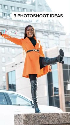 a woman in an orange coat and black boots standing on a ledge with her arms out