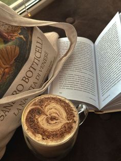 a cup of hot chocolate next to an open book and a bag on a table