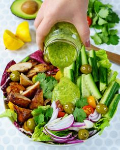 a person pouring dressing into a salad with lettuce, tomatoes, cucumber and olives