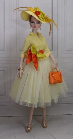 a doll wearing a yellow dress and hat with an orange purse in her hand, standing next to a white wall