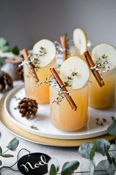 three glasses filled with drinks sitting on top of a white plate next to pine cones