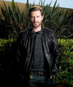 a man wearing a black leather jacket standing in front of some bushes and trees with his hands in his pockets