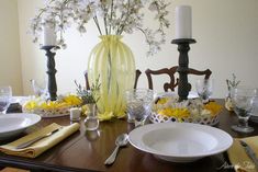 a dining room table is set with yellow and white flowers in vases, silverware, and candlesticks