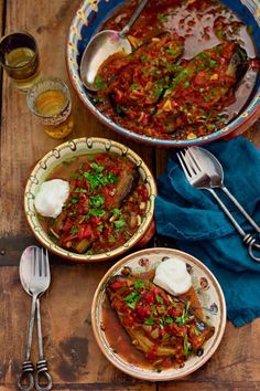 two bowls filled with stew next to silverware and forks on a wooden table top