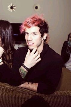 two people sitting on a couch with pink hair and piercings in their ears, one man is holding his hand up to the other woman's face