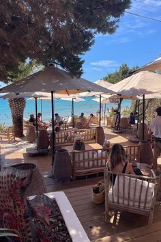 people are sitting at tables on the deck near the ocean with umbrellas over them