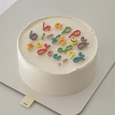 a white cake with colorful letters on it sitting on top of a table next to a piece of paper