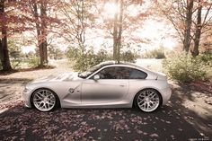 a silver sports car parked in front of trees with leaves on the ground and sun shining behind it