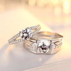 two wedding rings with diamonds on them sitting on a white surface, one has a diamond in the middle